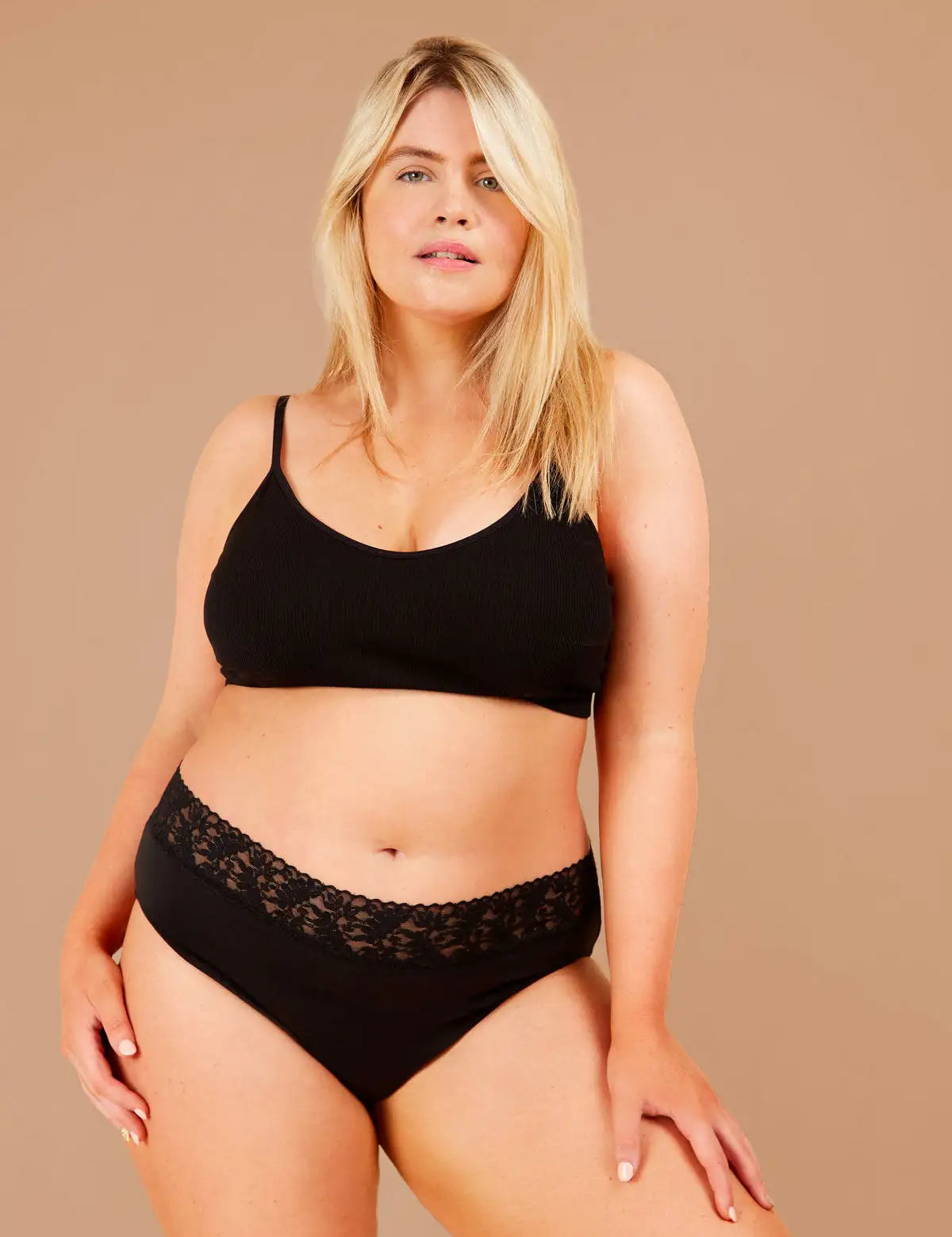 Blonde woman standing confidently, wearing black period pants with lacey floral trim gazes directly at the camera.